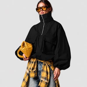 May include: A black long-sleeve pullover jacket with a high collar, silver front zipper, and two large chest pockets. The jacket is paired with blue denim jeans and a yellow and black plaid flannel shirt tied around the waist.