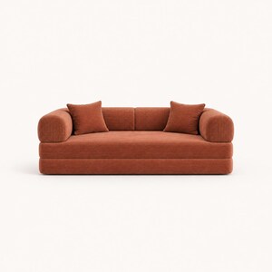 May include: A low-profile sofa upholstered in a rust-colored corduroy fabric. The piece features rounded armrests, a straight backrest, and two matching square throw pillows placed on the seat. The design has a horizontal ribbed texture.