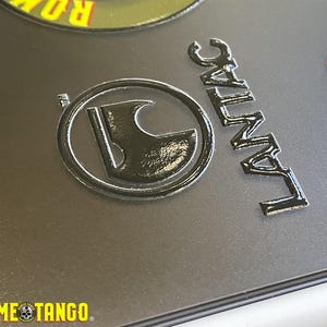 Puede incluir: Primer plano de emblemas y texto met&aacute;licos sobre una superficie gris. Los emblemas incluyen un dise&ntilde;o circular con un hacha estilizada y la palabra "LANTAC". El texto "ROME TANGO" tambi&eacute;n es visible.