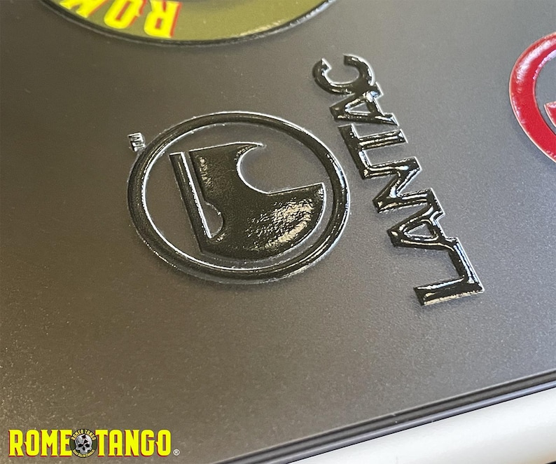 Puede incluir: Primer plano de emblemas y texto met&aacute;licos sobre una superficie gris. Los emblemas incluyen un dise&ntilde;o circular con un hacha estilizada y la palabra "LANTAC". El texto "ROME TANGO" tambi&eacute;n es visible.
