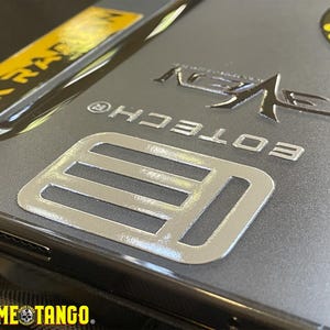 Puede incluir: Primer plano de una funda de tel&eacute;fono gris con detalles plateados y amarillos. La funda presenta el texto "EOTECH" y "ROME TANGO", junto con otros logotipos y dise&ntilde;os. La funda tiene un acabado met&aacute;lico.