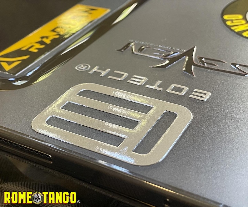 Puede incluir: Primer plano de una funda de tel&eacute;fono gris con detalles plateados y amarillos. La funda presenta el texto "EOTECH" y "ROME TANGO", junto con otros logotipos y dise&ntilde;os. La funda tiene un acabado met&aacute;lico.