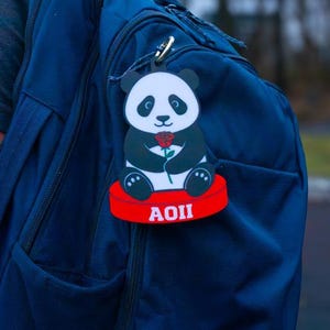 May include: A panda bear-shaped luggage tag with a red base and the letters AOII in white. The panda is black and white and holds a red rose. The tag is attached to a navy blue backpack.