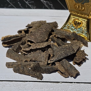 May include: A pile of dark brown agarwood chips, with a textured, layered appearance. The chips are scattered on a white surface, with a decorative gold and turquoise incense burner in the background. The wood has a natural, organic look.
