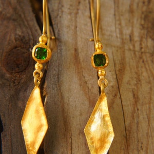 Könnte beinhalten: Ein Paar goldene Ohrringe mit grünen Edelstein-Akzenten. Die Ohrringe haben einen rautenförmigen Anhänger mit einer strukturierten Oberfläche.