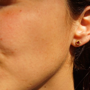 May include: A close-up of a person's ear with a small gold earring featuring a red gemstone.