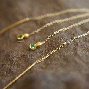 May include: A pair of gold earrings with green gemstone accents. The earrings feature a long chain with a small bar at the top and a green gemstone at the bottom.