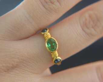 Könnte beinhalten: Ein goldener Ring mit einem ovalen grünen Edelstein in der Mitte, flankiert von zwei kleineren schwarzen Edelsteinen. Das Ringband ist mit Golddraht umwickelt, was ein strukturiertes Aussehen erzeugt. Schmuck.