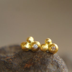 May include: A pair of gold earrings with small, round diamonds set in the center of each earring. The earrings are made of textured gold and have a cluster of three small spheres.
