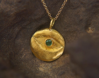 May include: Gold-toned pendant necklace featuring a circular charm with an eye design. The charm has a textured surface and a small, round, green gemstone in the center. The necklace chain is delicate and gold-colored.