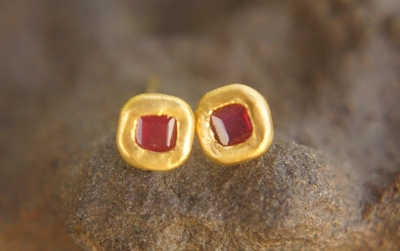 May include: A pair of gold earrings with small, square-shaped red gemstones set in the centre of each earring.
