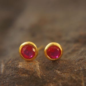 May include: A pair of gold stud earrings with vibrant red gemstones. The round gemstones are set in a gold bezel setting. The earrings are displayed on a textured, neutral-toned surface, highlighting the jewelry's color and design.