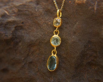 Può includere: Una collana con pendente color oro con tre pietre preziose. La pietra superiore è quadrata, quella centrale è rotonda e quella inferiore è ovale. Le pietre sono di colore azzurro e verde.