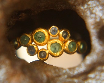 Könnte beinhalten: Ein goldener Ring mit einem Cluster aus grünen und blauen Edelsteinen. Der Ring ist in einer strukturierten Goldband gefasst.