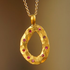 Puede incluir: Un collar con colgante dorado con un colgante texturizado en forma de lágrima. El colgante está adornado con pequeñas piedras preciosas rojas redondas. La cadena del collar también es dorada.