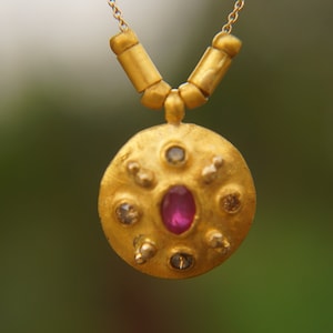 Puede incluir: Un collar de oro con un colgante redondo texturizado que presenta una piedra preciosa rosa rodeada de piedras blancas más pequeñas.
