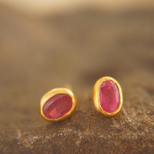 May include: A pair of gold-colored stud earrings, each featuring a vibrant red gemstone. The gemstones have an oval shape and are set in a smooth, polished gold bezel. The earrings are displayed on a textured, brown surface.
