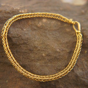 Könnte beinhalten: Ein goldfarbenes geflochtenes Armband. Das Armband besteht aus einer Reihe von ineinander verwobenen Strängen, wodurch ein strukturiertes Aussehen entsteht. Das Armband wird mit einem kleinen Verschluss gesichert. Das Armband wird auf einer strukturierten, braunen Oberfläche präsentiert.