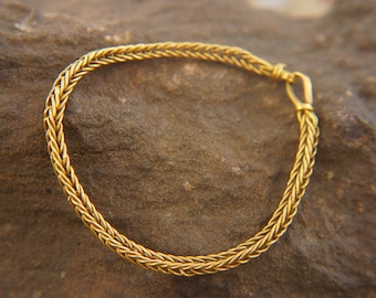 May include: A gold-colored braided bracelet. The bracelet is made of a series of interwoven strands, creating a textured appearance. The bracelet is secured with a small clasp. The bracelet is displayed on a textured, brown surface.
