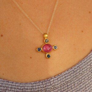 May include: A gold pendant necklace with a cross-shaped design. It features a large, round, pink gemstone at the centre, with four smaller blue gemstones. The necklace hangs from a fine gold chain.