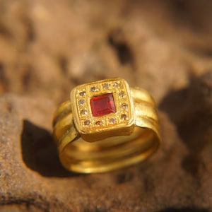 Op de afbeelding: Een gouden ring met een rode vierkante edelsteen omringd door kleinere heldere edelstenen. De ring heeft een dikke band met een getextureerd ontwerp.