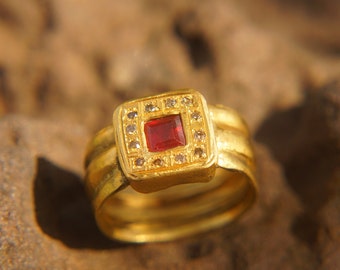 May include: A gold ring with a red square gemstone surrounded by smaller clear gemstones. The ring has a thick band with a textured design.