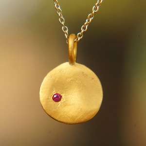 Op de afbeelding: Een gouden ketting met een ronde hanger met een klein rood steentje.