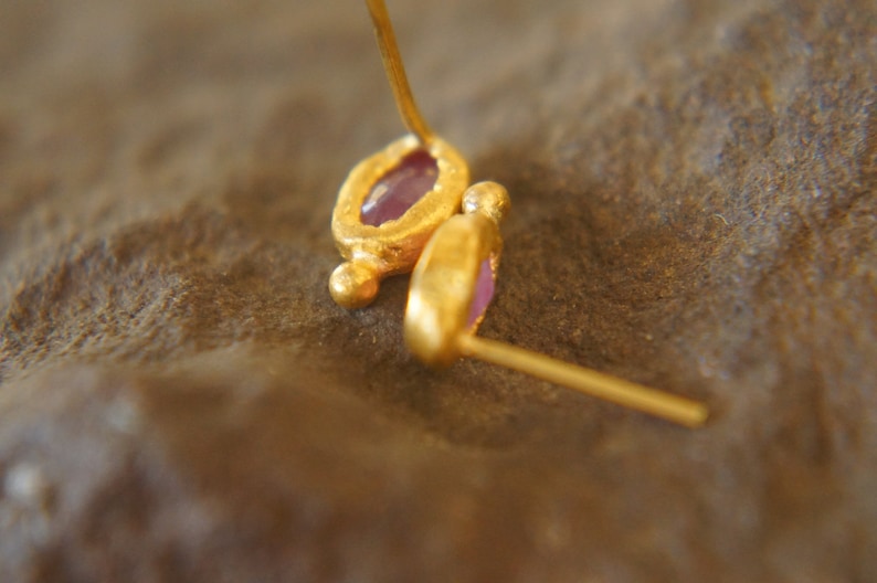Puede incluir: Un par de pendientes de oro con peque&ntilde;as piedras preciosas p&uacute;rpuras. Los pendientes est&aacute;n hechos de metal texturizado y tienen un dise&ntilde;o sencillo.
