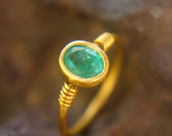 May include: A gold ring with an oval, emerald-green gemstone. The ring's band is a simple, thin gold, with a coiled design near the gemstone. The gemstone is set in a gold bezel.