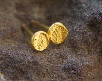 May include: A pair of gold-colored stud earrings. Each earring features a circular design with a leaf imprint. The earrings are set against a textured, dark gray surface.