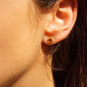 May include: A close-up of a person's ear with a small gold earring featuring a red gemstone.