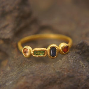 May include: A gold ring with four colorful gemstones. The ring features a peach-colored stone, a green rectangular stone, a dark blue oval stone, and a red stone. The ring is set on a textured surface.