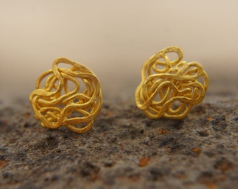 May include: A pair of gold-colored earrings with a knotted, twisted design. The earrings are small and delicate, and they are set against a textured, brown background.