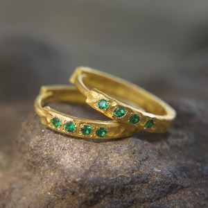Puede incluir: Un par de pendientes de aro de oro con textura martillada y una fila de pequeñas piedras preciosas verdes.