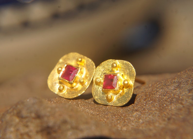 May include: A pair of gold earrings with a square red gemstone in the centre of each earring. The earrings have a textured gold finish and are set in a square shape.