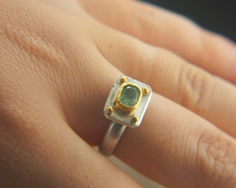 May include: A silver ring with a square, two-tone setting. The setting features a gold border around a green gemstone and is accented with small gold details. The ring is on a finger.