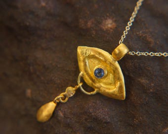 May include: A gold-colored pendant necklace with a leaf-shaped charm. The charm features a small, round, blue gemstone in the center and a dangling teardrop-shaped element. The necklace chain is delicate and gold-toned.