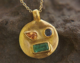 May include: A gold pendant necklace with a round charm featuring a face with three gemstones: a green emerald, an orange sapphire, and a blue sapphire.