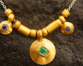 May include: A gold necklace with a large round pendant featuring a green emerald gemstone. The necklace also has smaller round charms with blue sapphires.