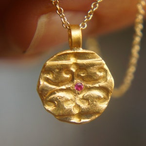 Può includere: Una collana con un ciondolo rotondo in oro decorato con una piccola pietra rossa al centro. Il ciondolo ha un design testurizzato e dettagliato.