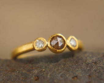 Puede incluir: Un anillo de oro con tres diamantes engastados en bisel. El diamante central es marrón, y los dos diamantes laterales son blancos.