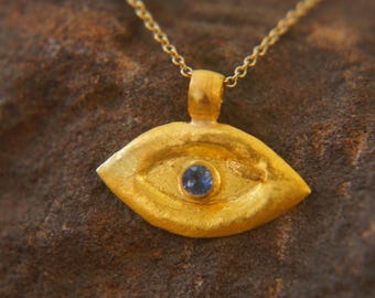 May include: A gold-colored pendant necklace featuring an eye-shaped charm. The charm has a textured surface and a small, round, blue gemstone in the center. The necklace chain is also gold-colored.