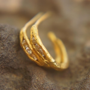 May include: A pair of gold-colored hoop earrings. Each earring features a textured, organic design and is embellished with small, sparkling gemstones. The earrings are displayed against a neutral, textured background.