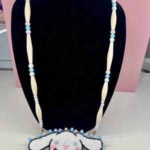 May include: A beaded necklace with a white cartoon character pendant, featuring black and pink details. The necklace has a cream-coloured beaded chain with light blue and white accents. The pendant is surrounded by light blue and white beads.