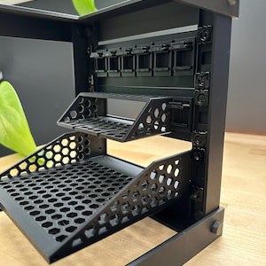 May include: A black metal rack with multiple shelves and a honeycomb pattern. The rack has a rectangular frame and is designed to hold various items. The shelves are angled and appear to be made of a durable material.