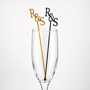 May include: Two thin drink stirrers placed inside a clear glass flute. One stirrer is gold and the other is black. Both stirrers feature a decorative topper with the letters R and S connected by an ampersand symbol in a serif font style.