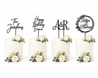 May include: Four black cake toppers with custom text including The Jardieus, Happy Birthday Emma, A&R, and Katya & Andrew. Each topper is displayed on a white, horizontally ribbed cake decorated with white roses and small green leaves.