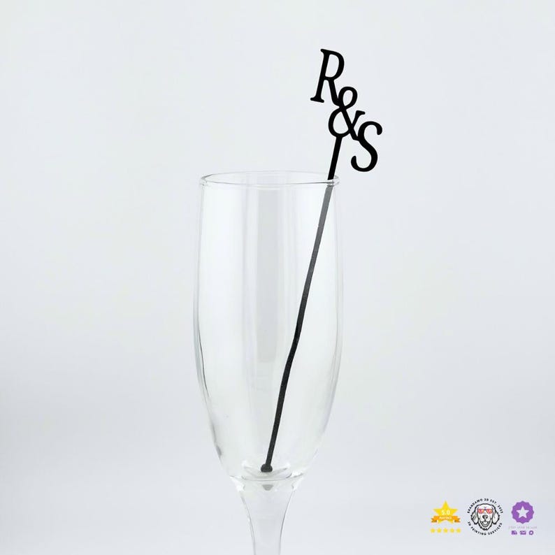 May include: A clear glass champagne flute containing a black drink stirrer. The stirrer features a thin vertical stem topped with the black cutout letters R and S separated by an ampersand symbol, creating a decorative monogram topper.