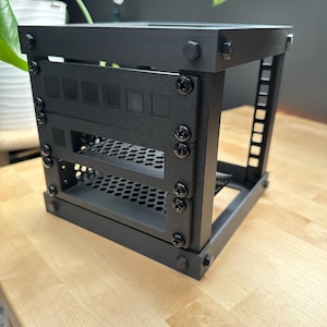 May include: A black metal computer server rack with multiple mounting points and a perforated shelf. The rack is assembled with black screws and bolts, and is designed to hold computer components. The rack is sitting on a light-coloured wooden surface.