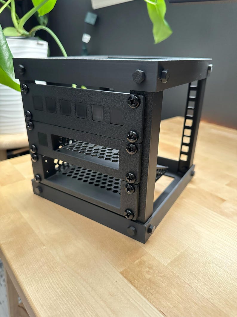May include: A black metal computer server rack with multiple mounting points and a perforated shelf. The rack is assembled with black screws and bolts, and is designed to hold computer components. The rack is sitting on a light-coloured wooden surface.